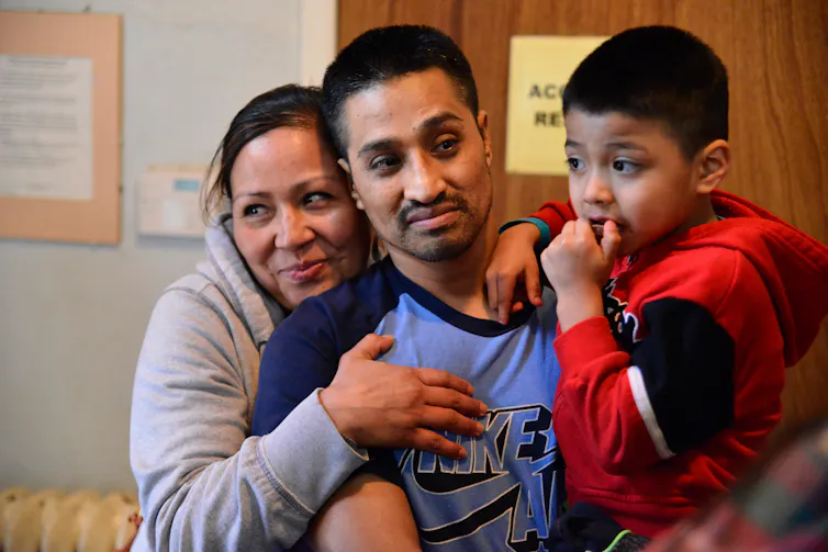 Man in blue shirt holds a child as woman hugs him from behind
