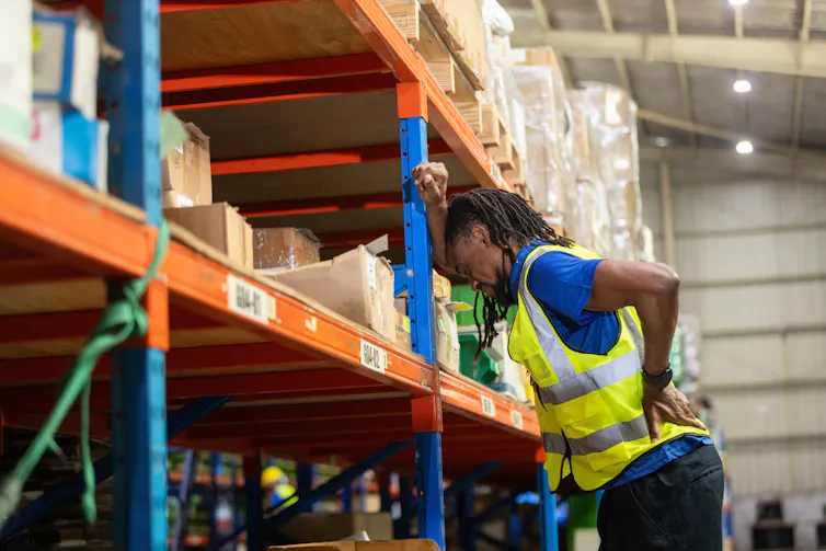 male warehouse worker leaning on shelves and grabbing his back in pain.