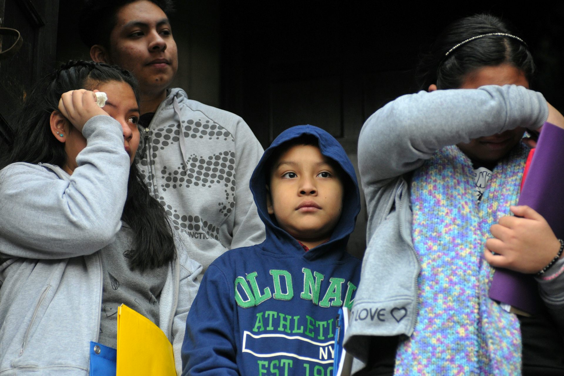Four children of various ages stand together, two of them wiping face with hand or arm
