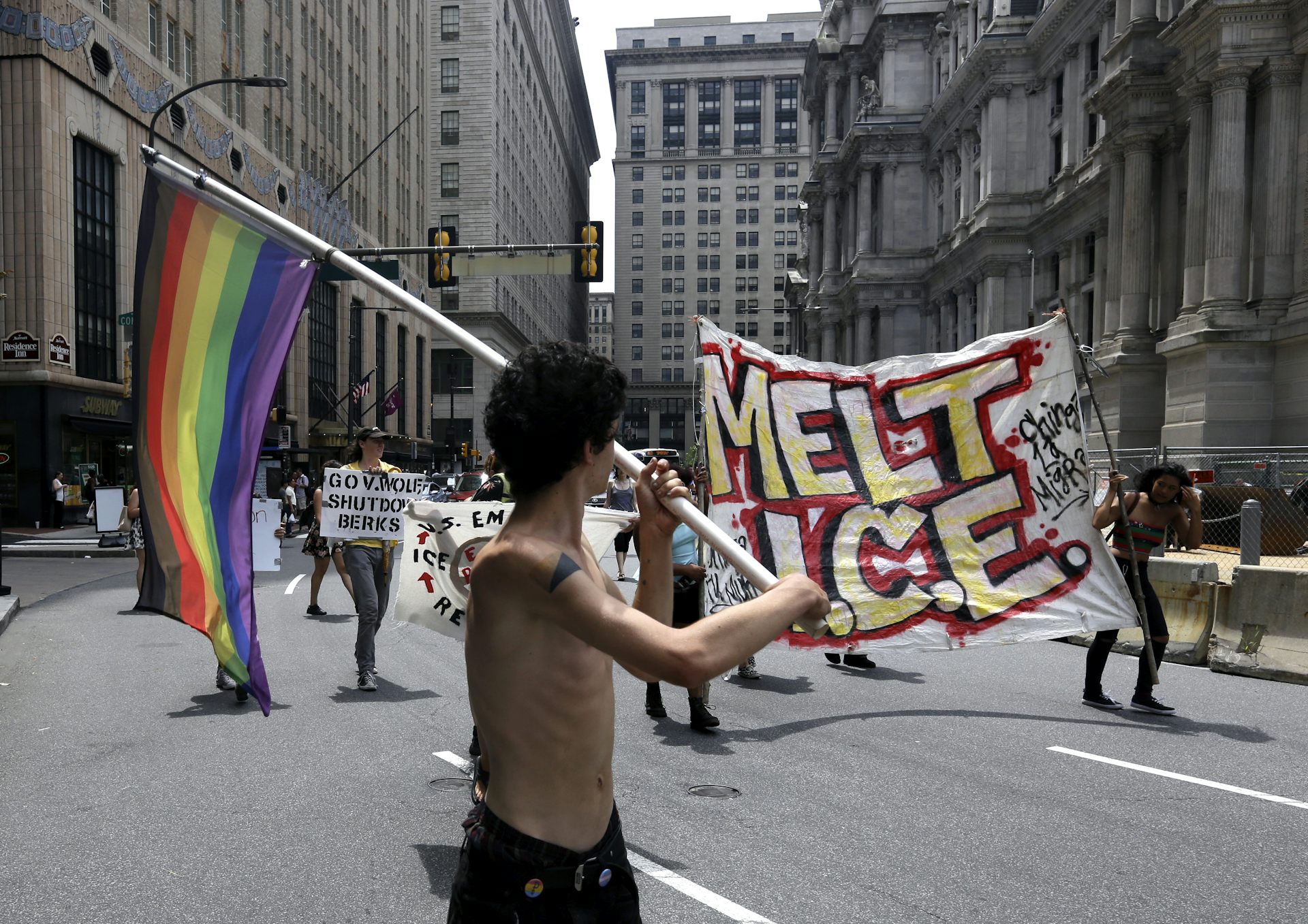 Young shirtless man holds rainbow flag while protesters behind him carry banner that says 'Melt ICE'