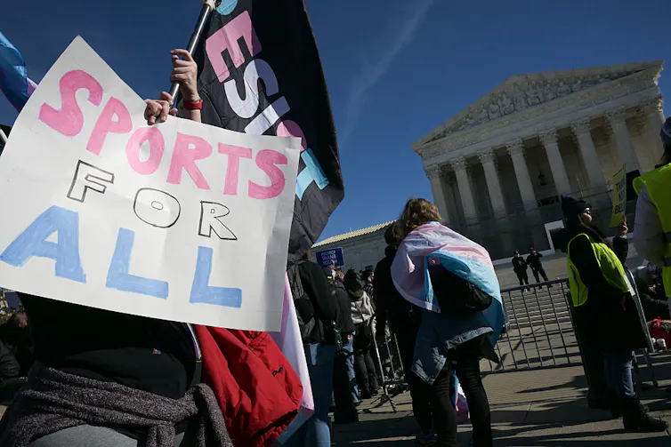 Person holding a sign reading 'SPORTS FOR ALL' in front of U.S. Supreme Court; other people bearing trans flags