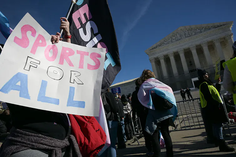 Person holding a sign reading 'SPORTS FOR ALL' in front of U.S. Supreme Court; other people bearing trans flags