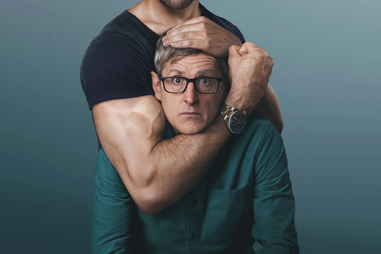 A middle-aged man in glasses stares at the camera as a muscular arm grips his head and clenches a fist.