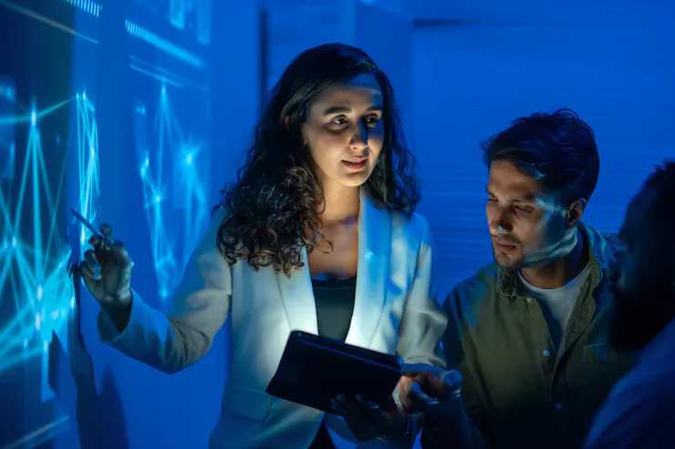 A woman wearing a white jacket points to a programming screen while other members of the team look on.