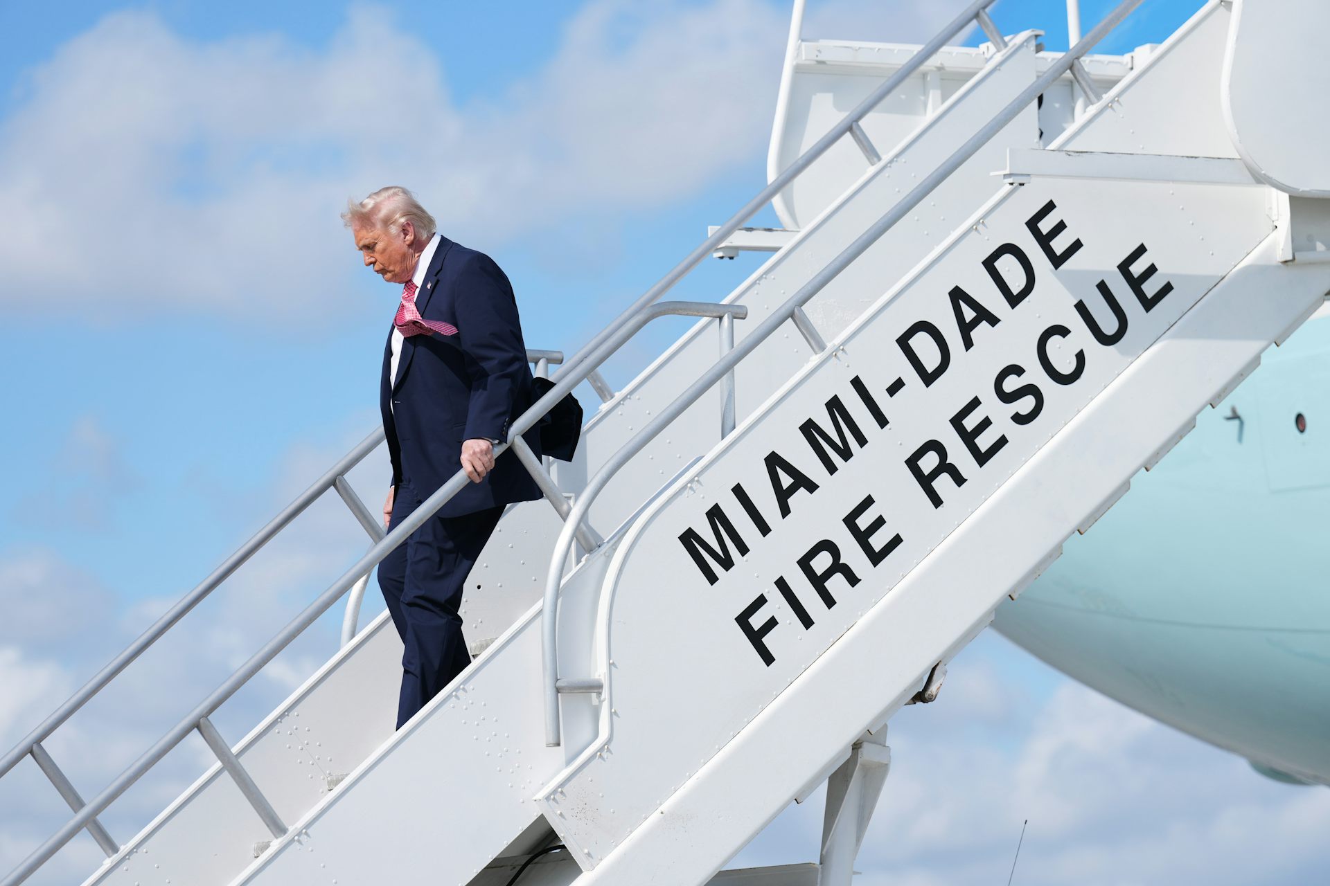 A rotund man with blond-ish white fluffy hair blowing in the breeze disembarks from an aircraft down a set of stairs that reads miami-dade fire rescue