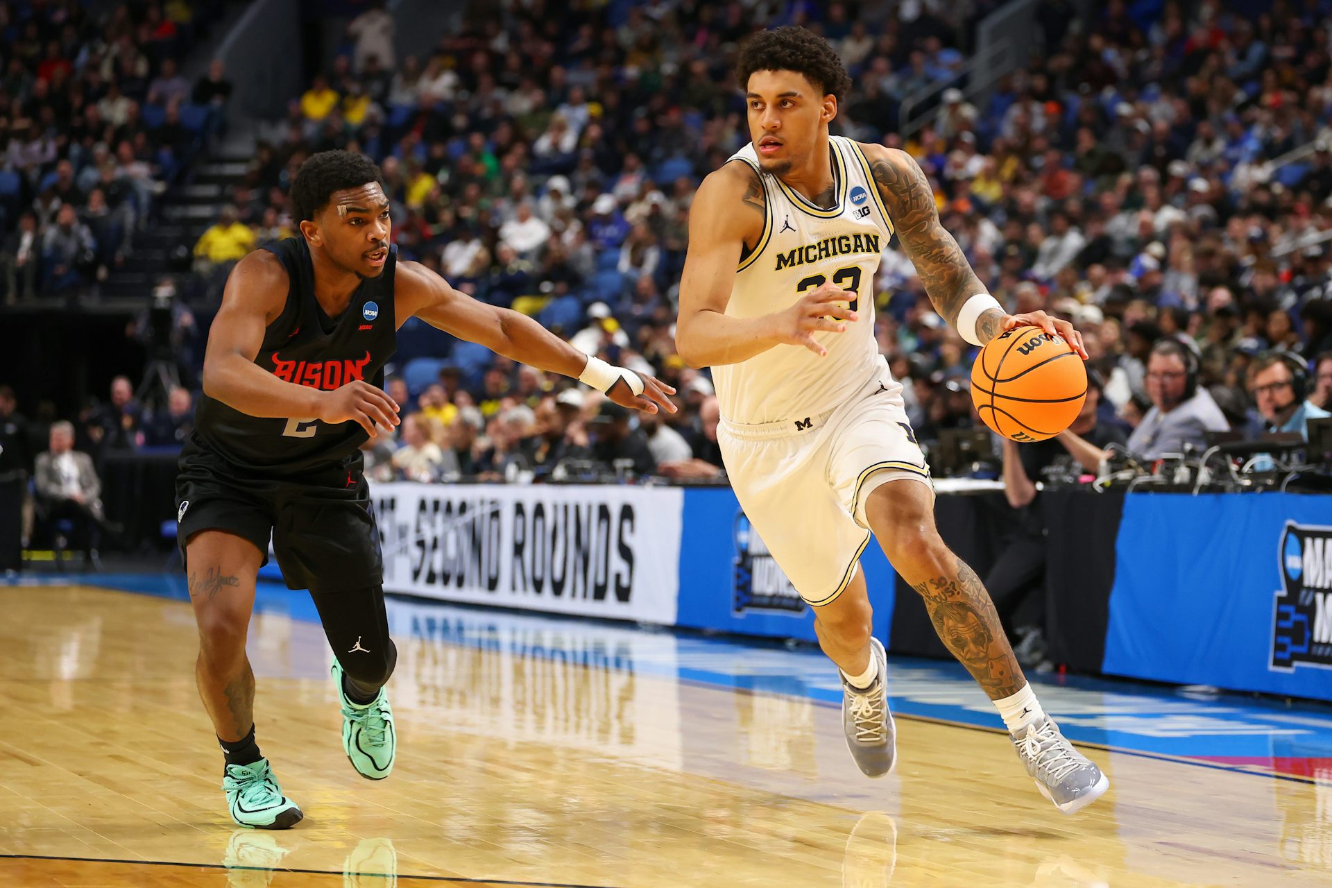 Young man in white jersey dribbles basketball while young man wearing black jersey guards him.
