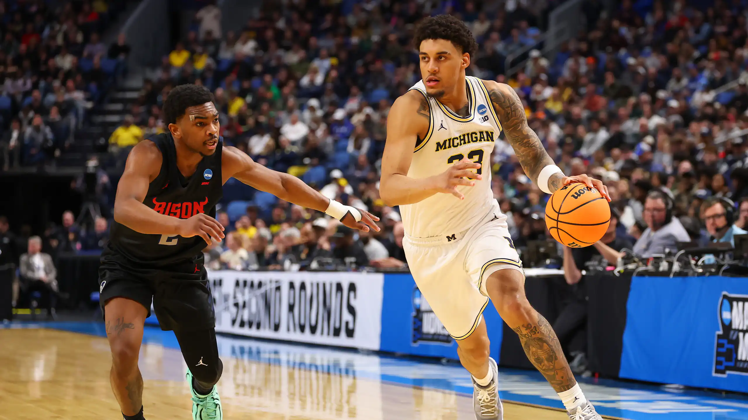 Young man in white jersey dribbles basketball while young man wearing black jersey guards him.