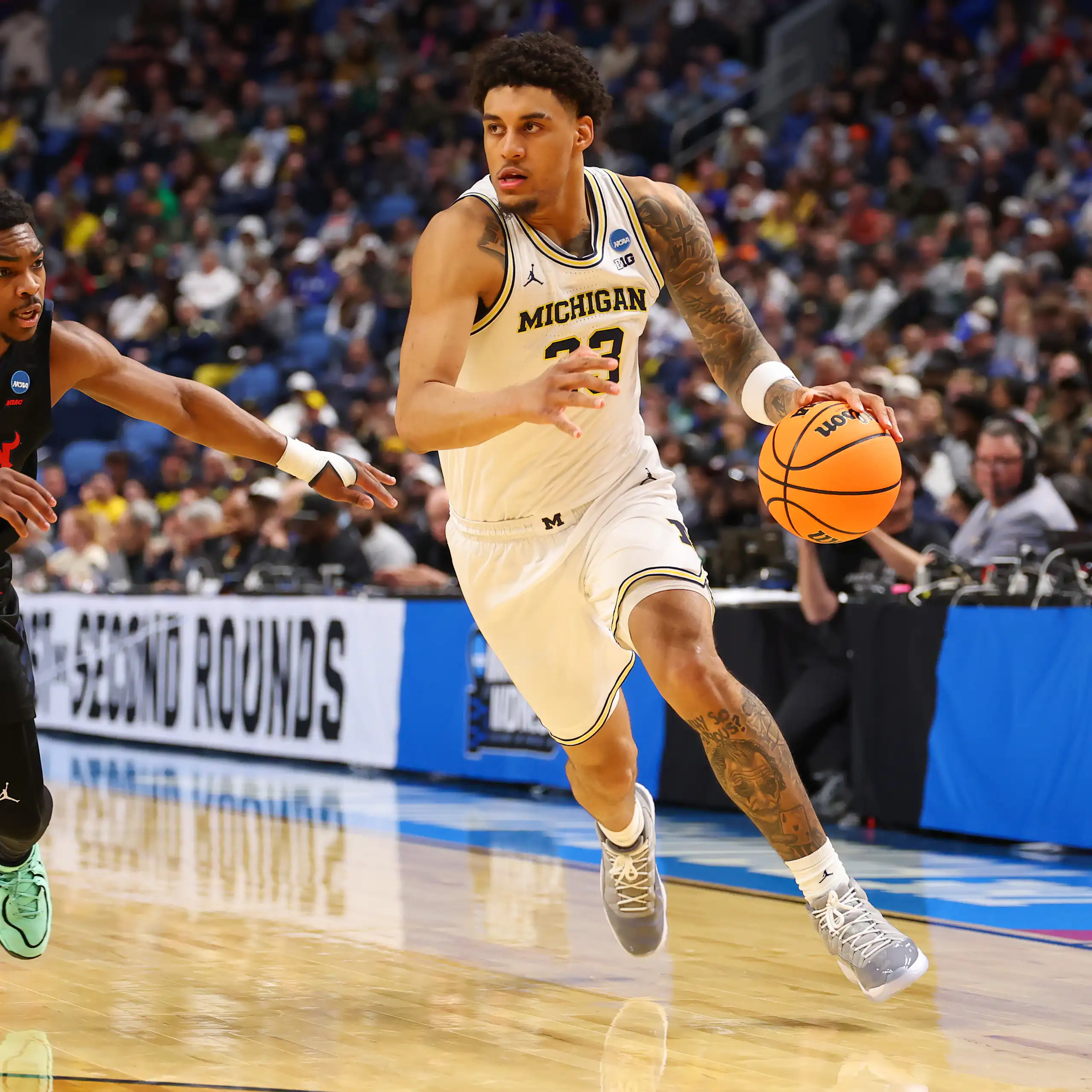 Young man in white jersey dribbles basketball while young man wearing black jersey guards him.