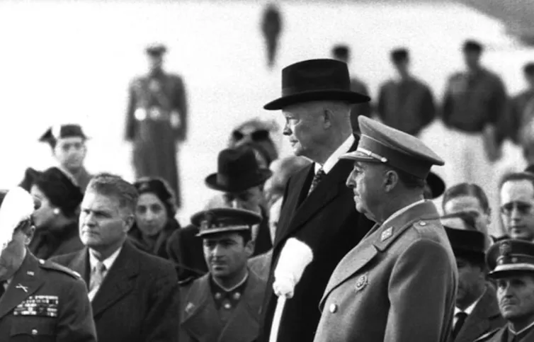 Dos hombres, un militar y uno de traje, esperan de pie rodeados de más hombres.