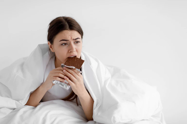 Woman eats chocolate while wrapped in bedsheets.