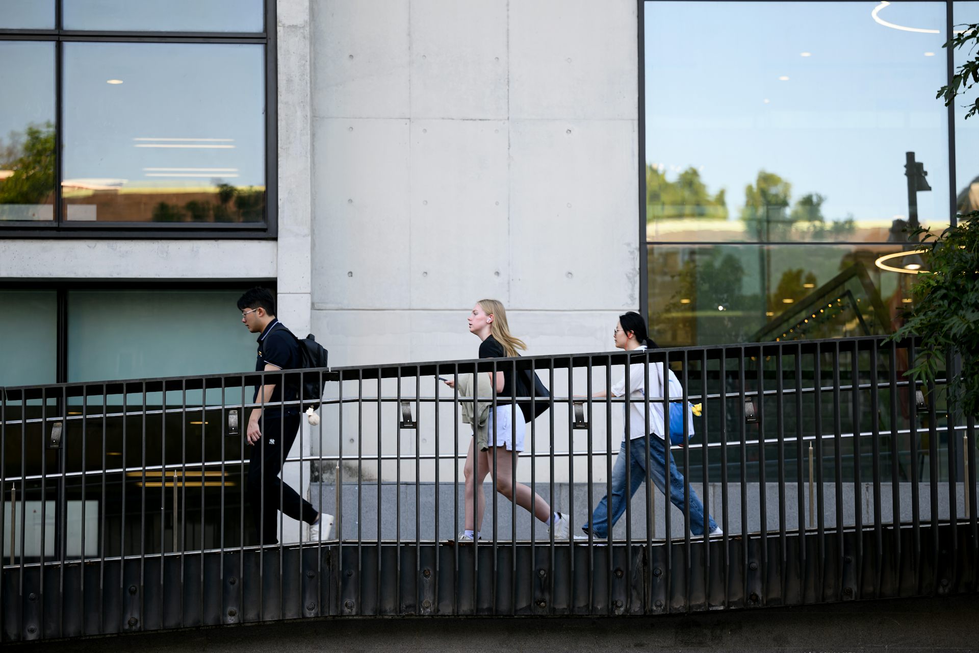 Three young people walk across a footbridge, in front of a tall building with large glass windows. 