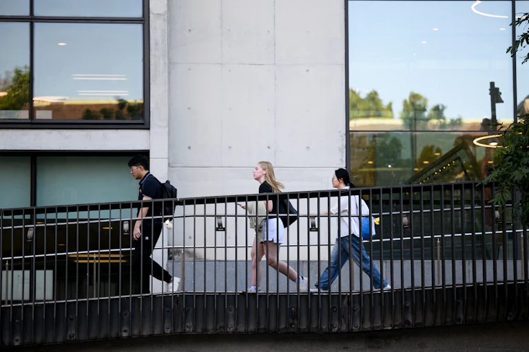 Three young people walk across a footbridge, in front of a tall building with large glass windows.