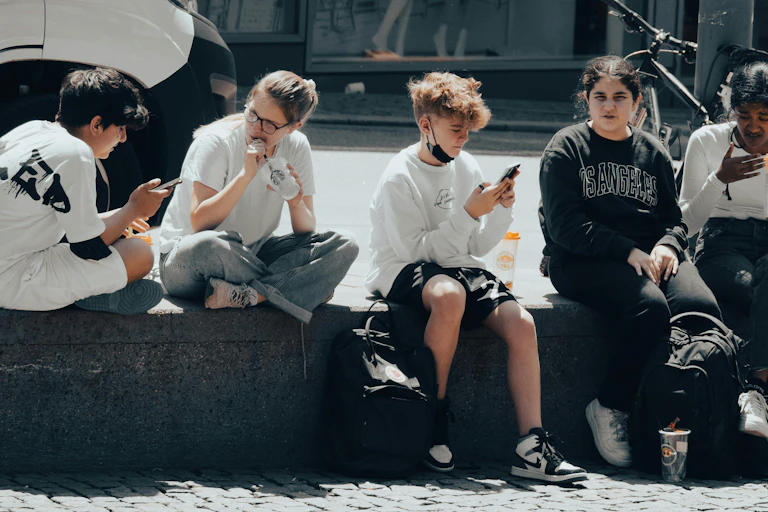 A group of teenagers sitting on the street, looking at their phones and talking.