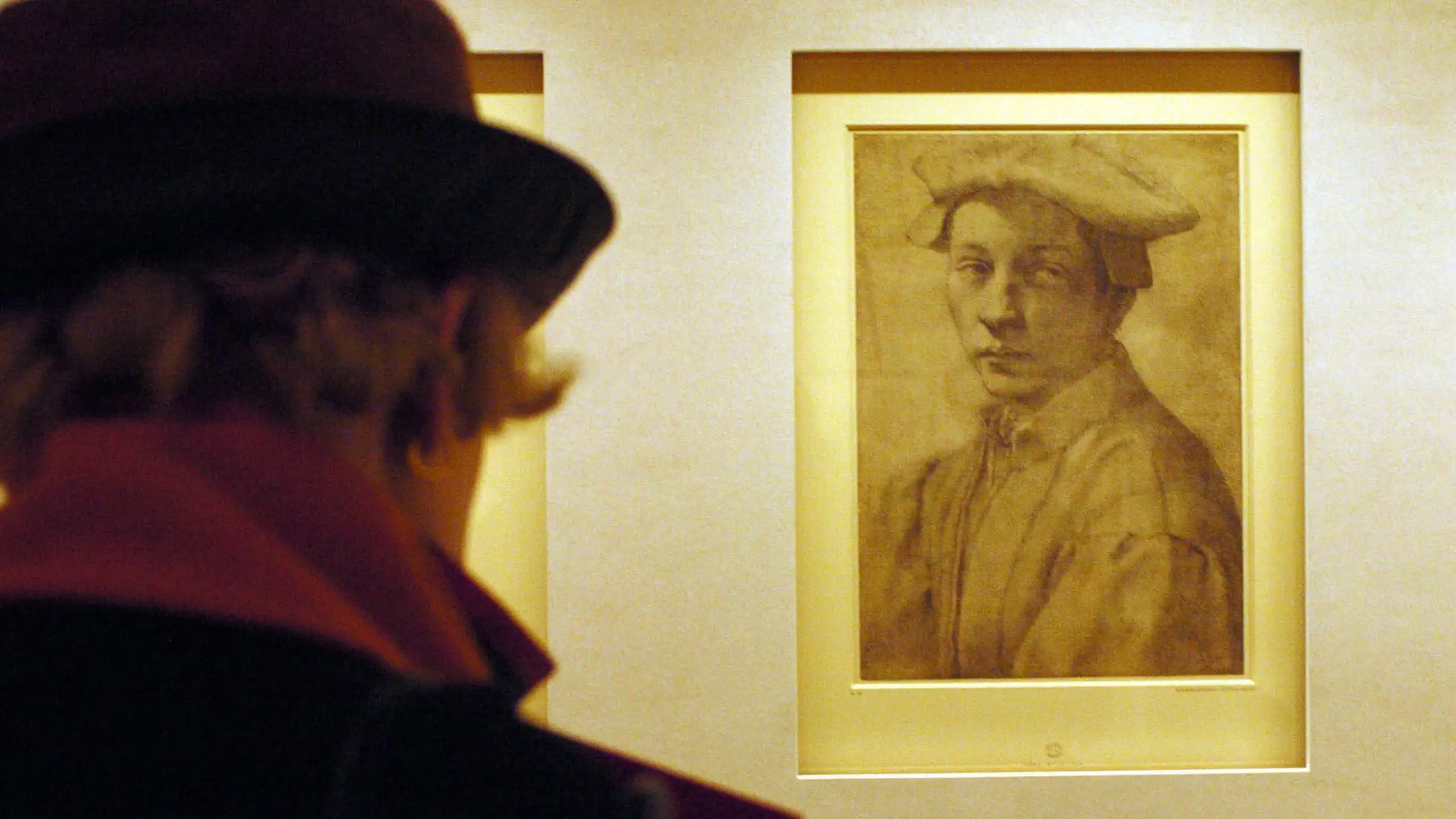 A woman in a museum gallery views of sketched portrait of a young man wearing a hat.