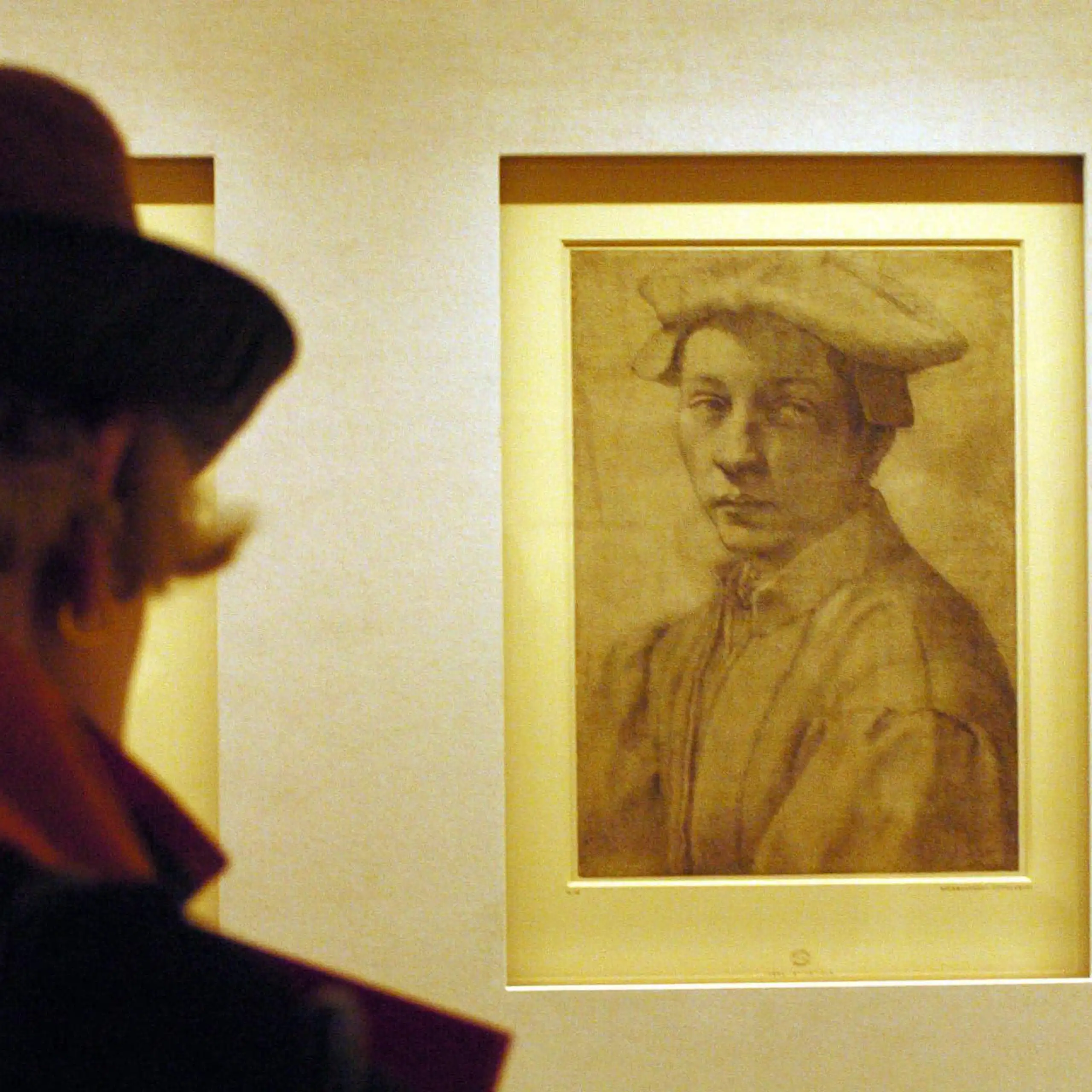 A woman in a museum gallery views of sketched portrait of a young man wearing a hat.