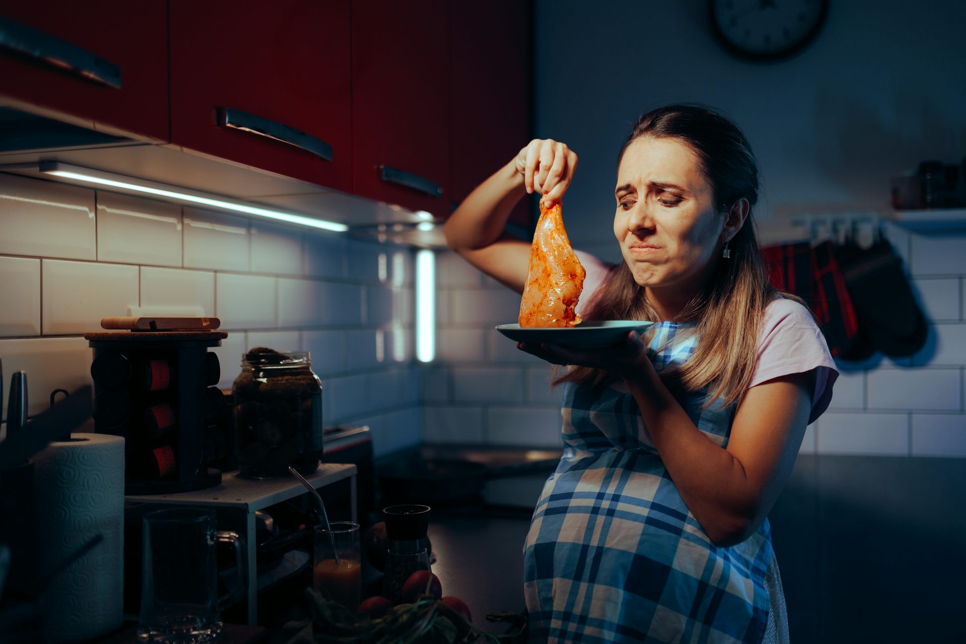 A pregnant woman standing next to a stove holds up a raw, seasoned chicken breast. She looks at it in disgust.