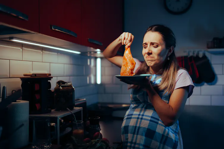 A pregnant woman standing next to a stove holds up a raw, seasoned chicken breast. She looks at it in disgust.