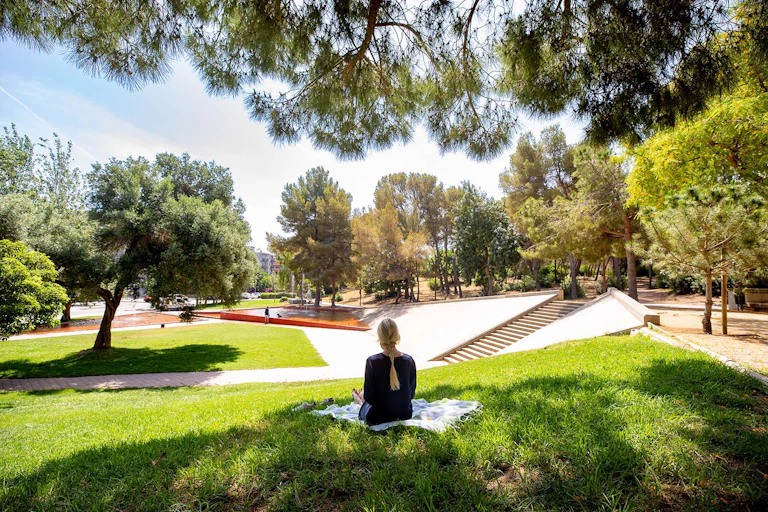 Una mujer está sentada en la hierba de espaldas en un parque con pinos y una fuente de agua al fondo