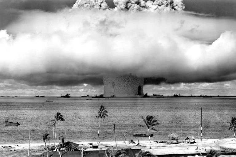 A large mushroom cloud spreads above a beach and water.