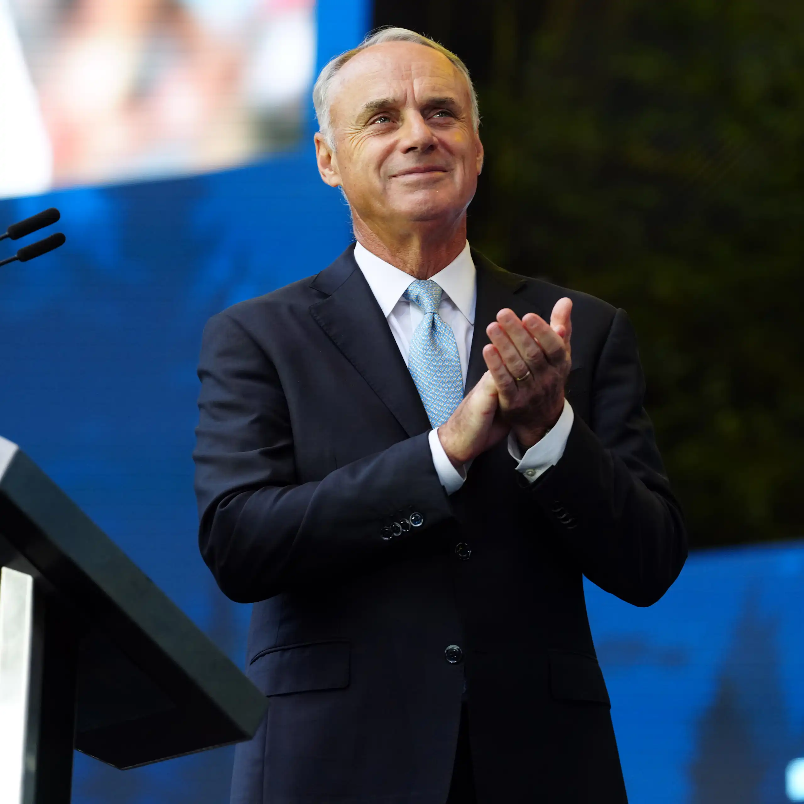 Man wearing suit grins and claps while standing next to a podium.