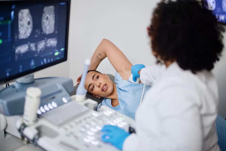 Patient lying on side with arm raised, health care provider holding ultrasound probe on the side of their breast, images displayed on the machine's screen