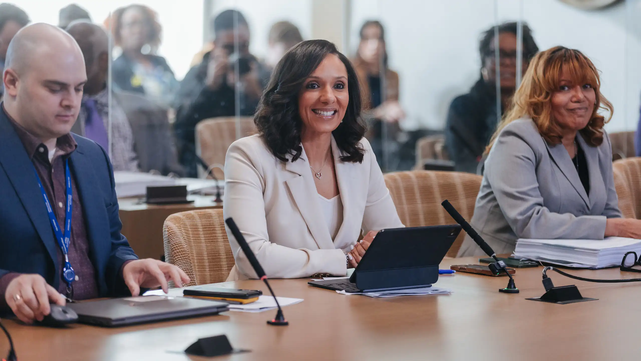 A bright-eyed Black woman wearing a light-colored suit sits at a conference table with a laptop in front of her. Aides flank her.
