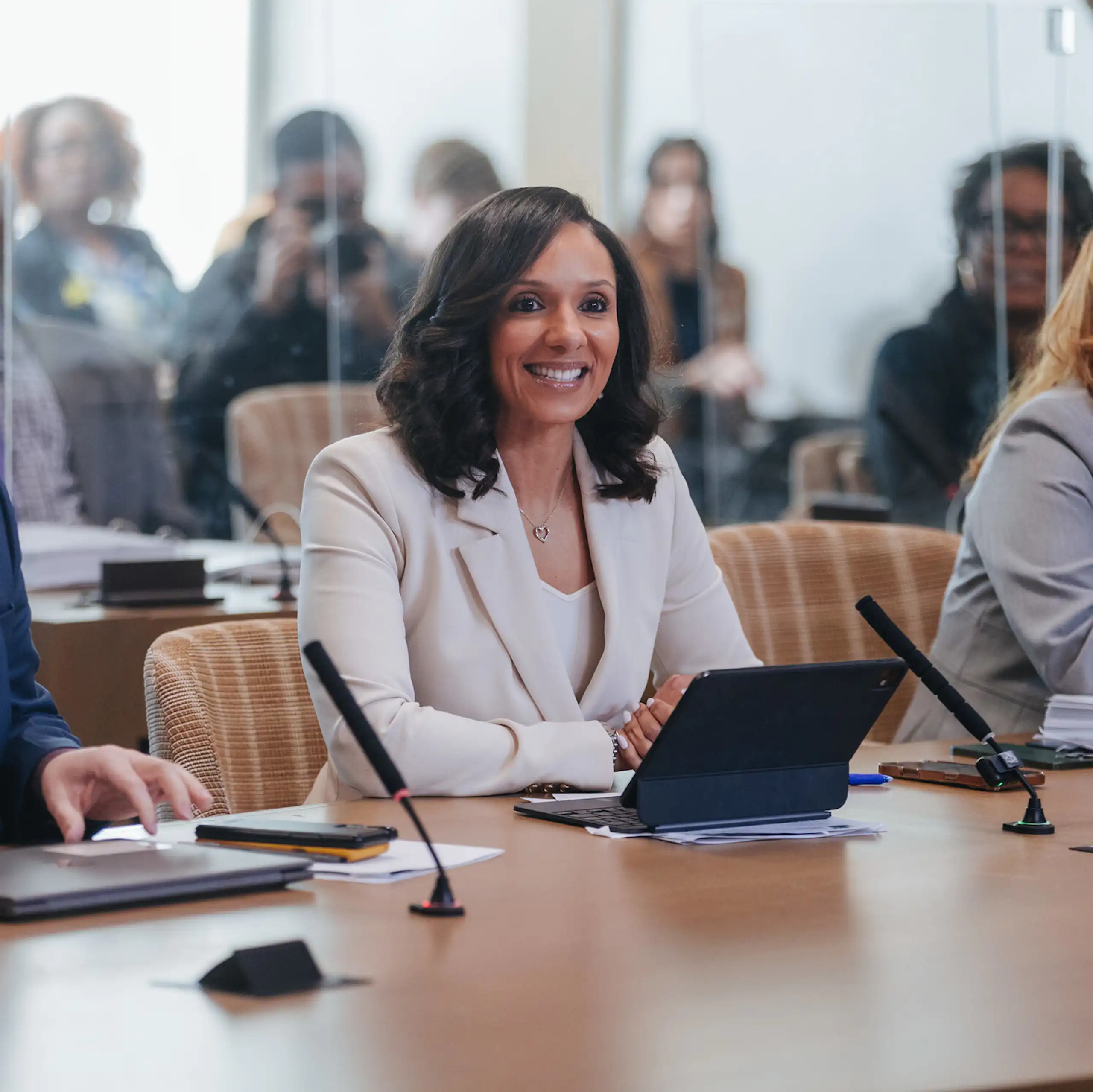 A bright-eyed Black woman wearing a light-colored suit sits at a conference table with a laptop in front of her. Aides flank her.