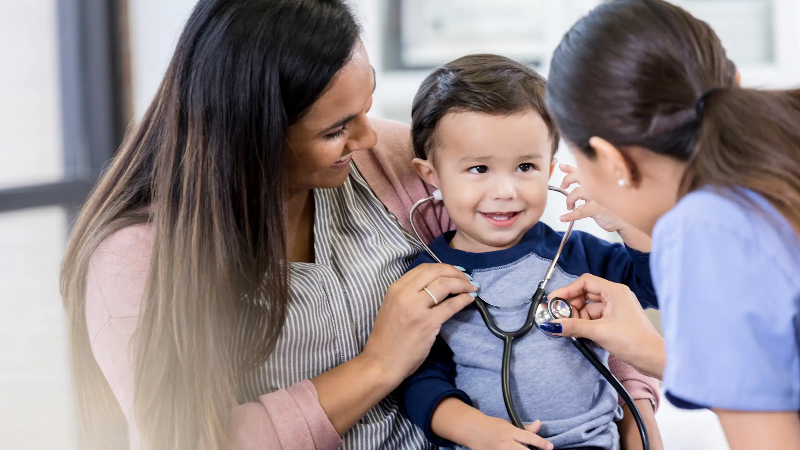 Health care provider and mom help toddler try on a stethoscope