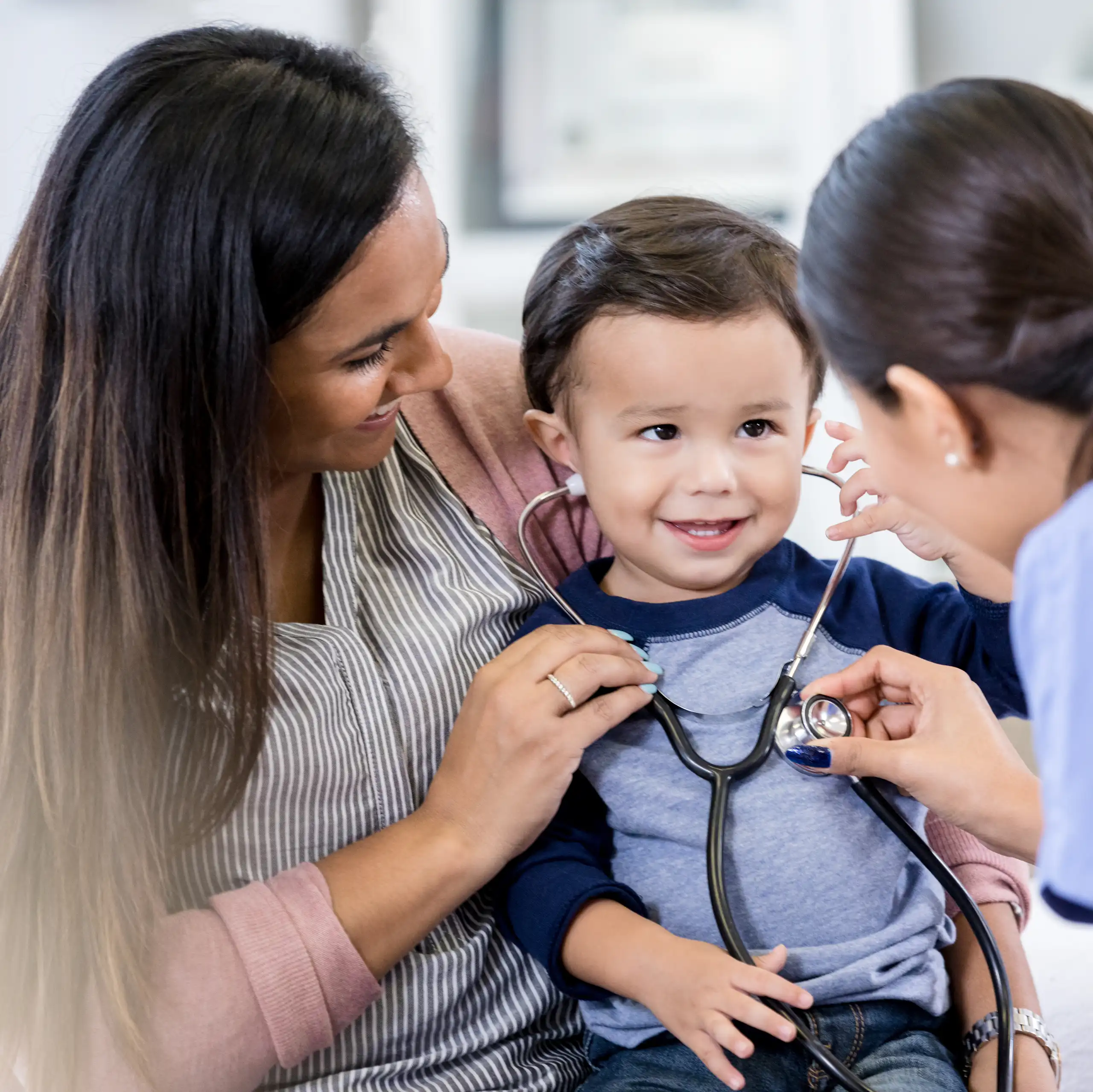 Health care provider and mom help toddler try on a stethoscope