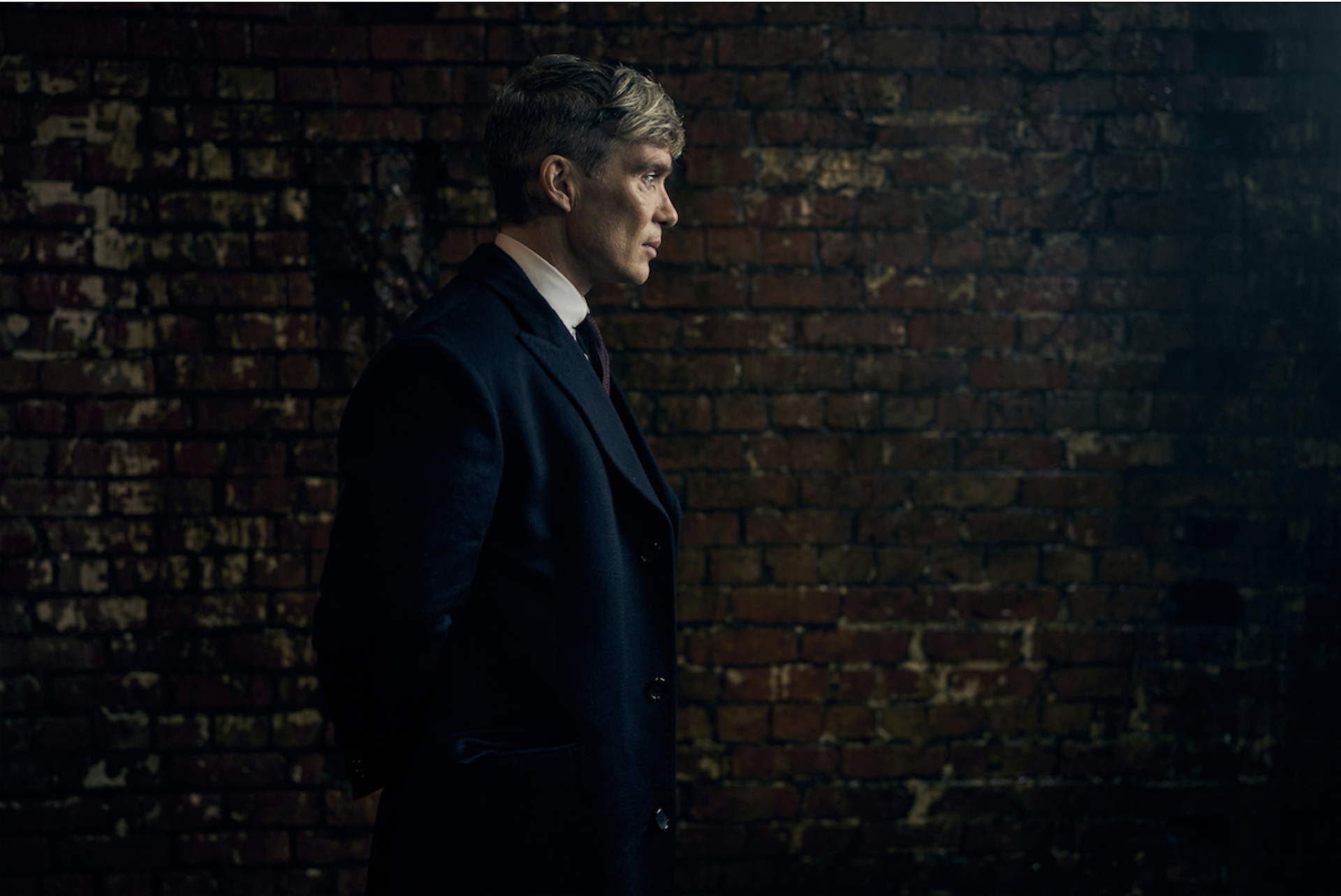 A man standing in profile looking pensive in a dark suit against a dark shadowy background.