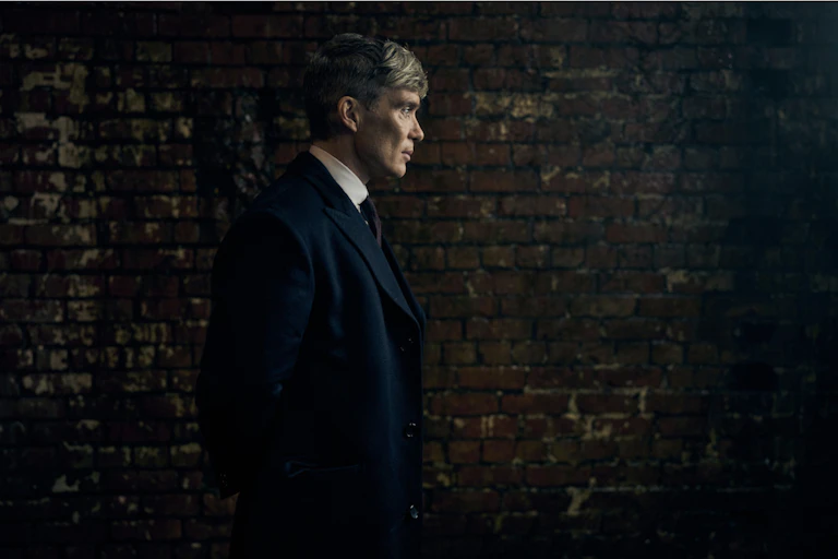 A man standing in profile looking pensive in a dark suit against a dark shadowy background.
