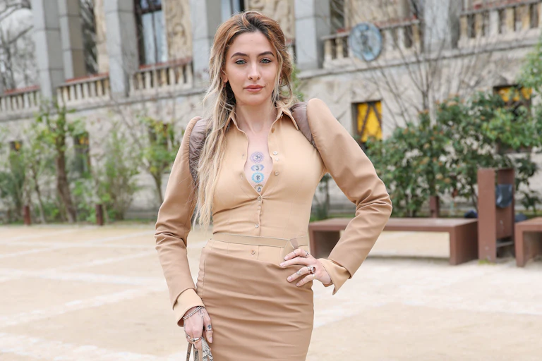 A woman in a beige outfit and long hair poses in a European setting.