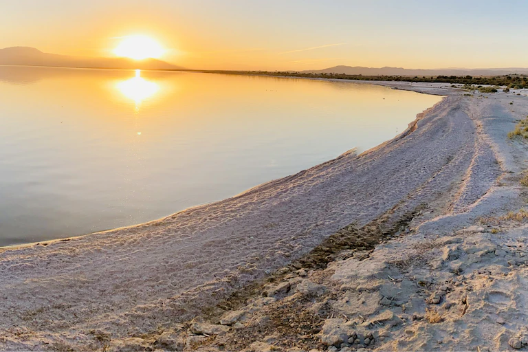 A sandy shoreline with a large lake in the background and sun at the horizon