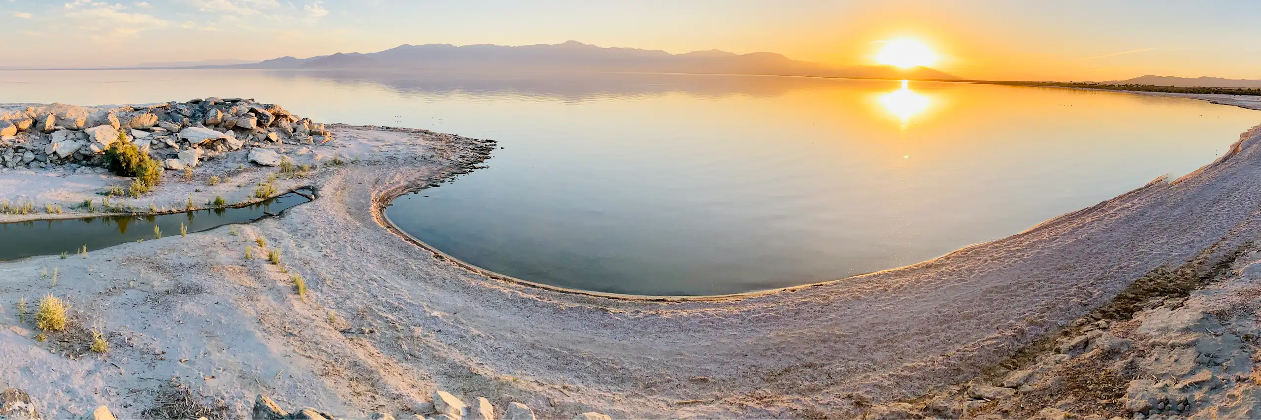 A sandy shoreline with a large lake in the background and sun at the horizon