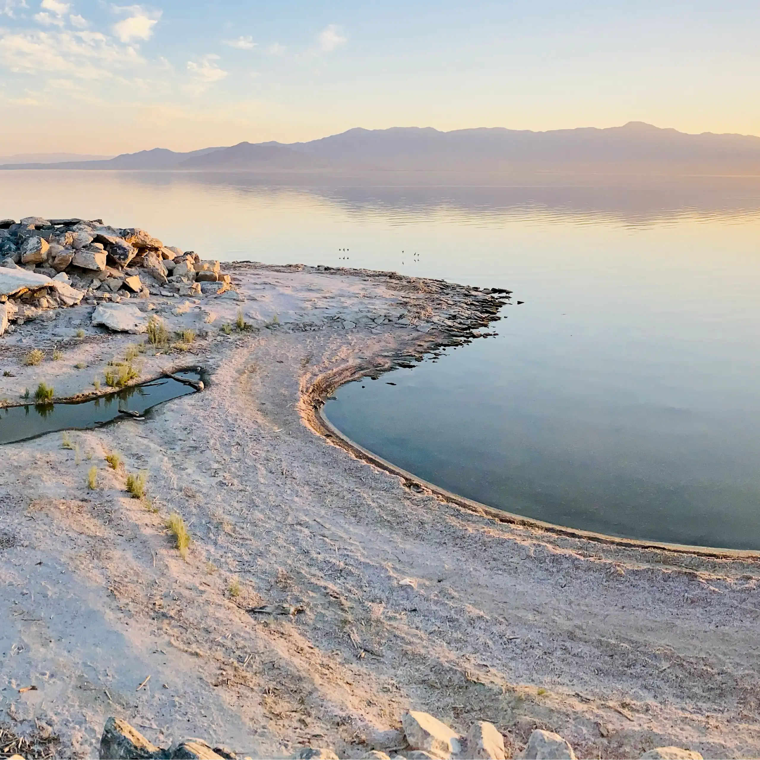 A sandy shoreline with a large lake in the background and sun at the horizon