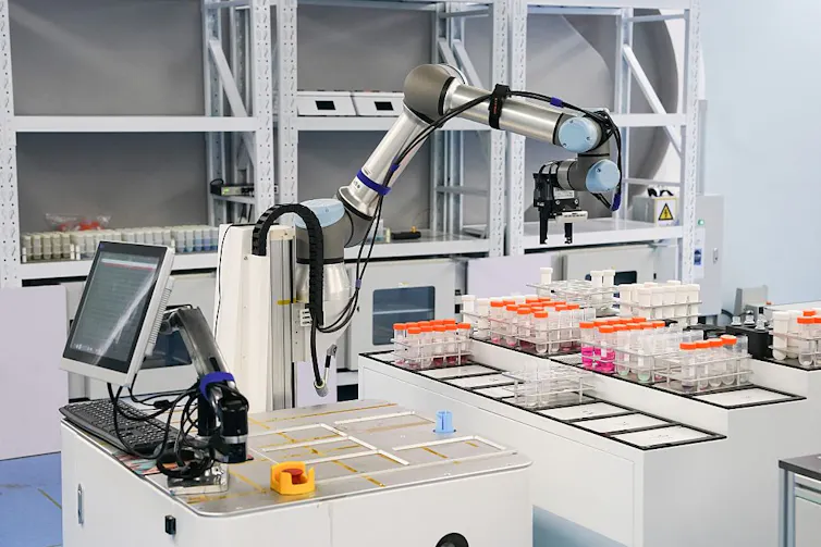 Robotic arm hovering over trays of specimen containers in a lab, a computer monitor behind it