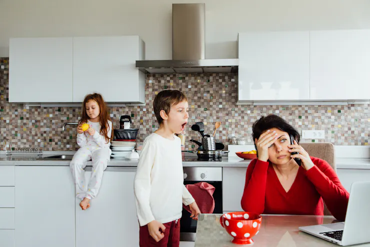 Middle-aged woman looking overwhelmed sitting at a kitchen table with two kids in the background.