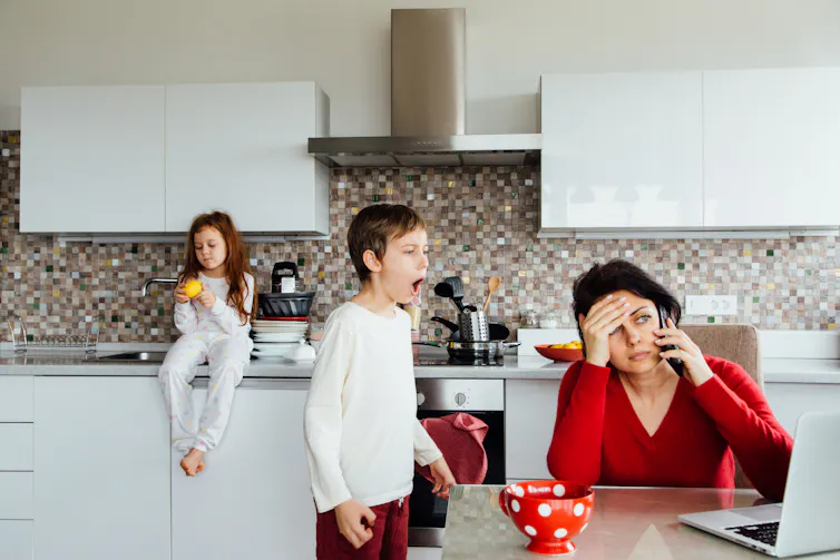 Middle-aged woman looking overwhelmed sitting at a kitchen table with two kids in the background.