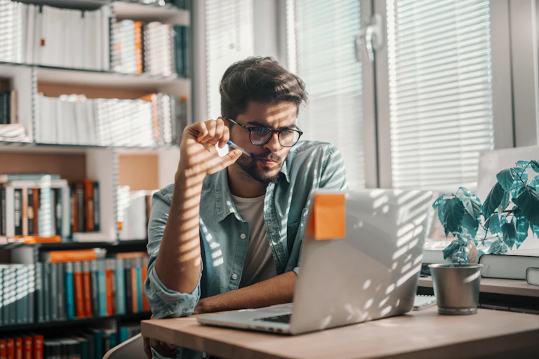 A young man looks at a computer screen. There is a bookshelf behind him. Filtered light comes through blinds to his side.