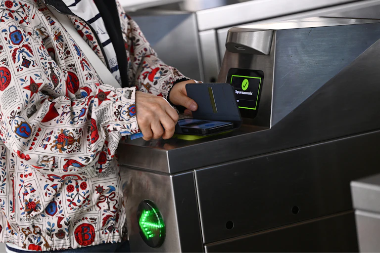 A woman's hands holds a phone over a ticket scanner at a train station turnstile.