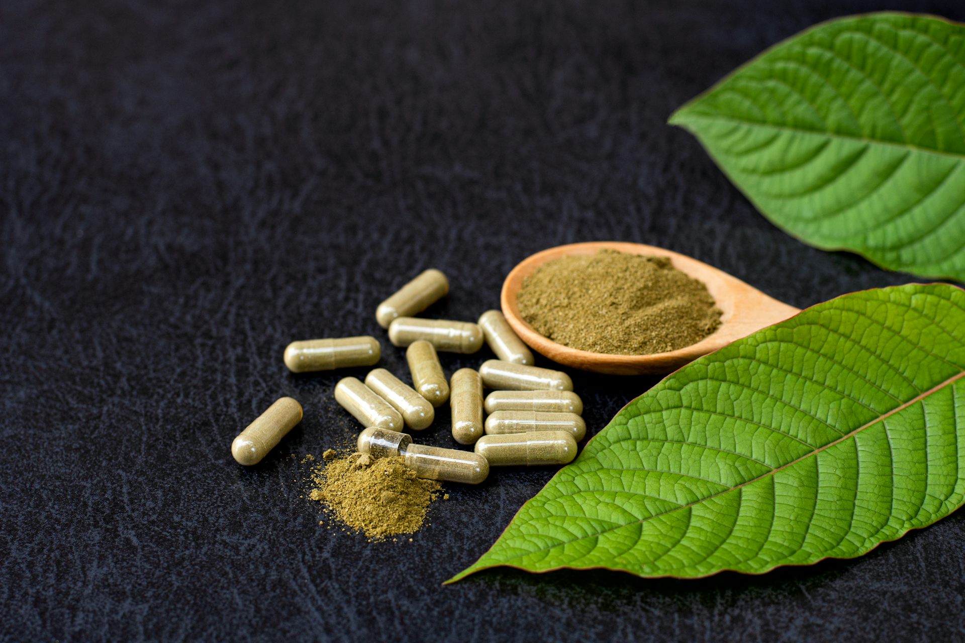 Brownish-green powder sitting on a spoon aside capsules containing the same powder and leaves of a plant off to the side.