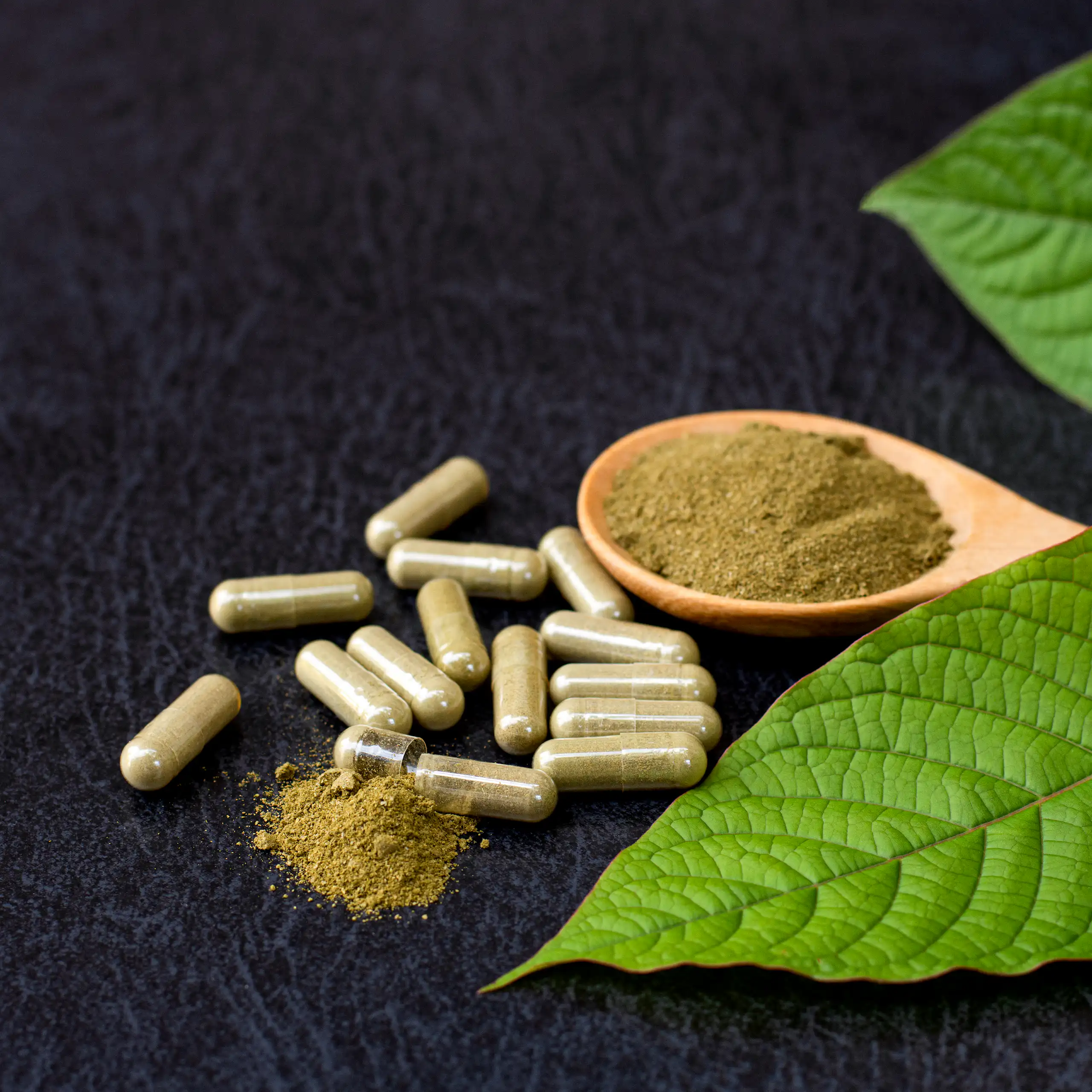 Brownish-green powder sitting on a spoon aside capsules containing the same powder and leaves of a plant off to the side.