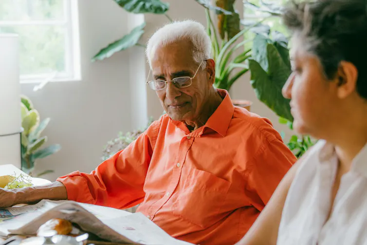 A man with grey hair in an orange shirt sitting a table with a woman out of focus and plants in the background