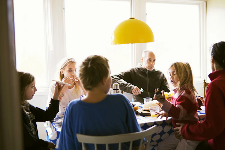 A family group sits at a table having a meal. A young child stands a man wears a serious expression.