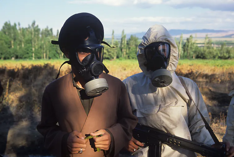 Two people stand in a field wearing gas masks.