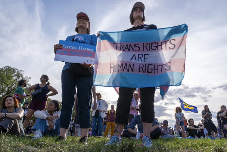 Dos mujeres paradas en un campo durante una marcha sosteniendo una bandera trans en apoyo de los derechos trans.