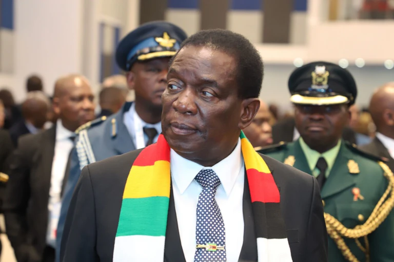 A man wearing a suit and scarf walking ahead of a crowd of people who include armed men