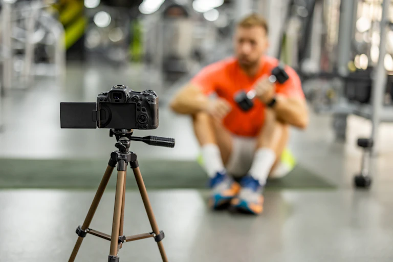 A blurry image of a man in an orange shirt lifting a weight as he sits on the floor in front of a camera tripod.
