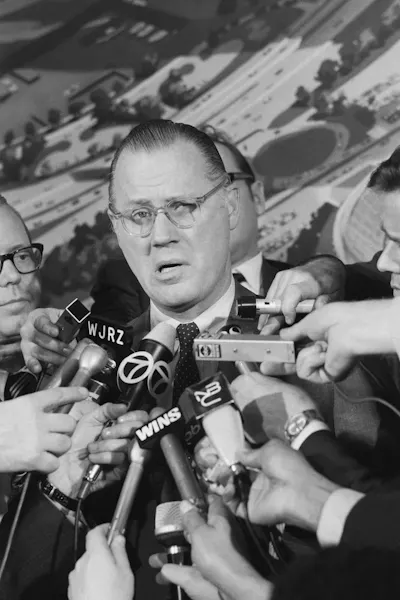 Black-and-white photo of be-speckled man speaking into reporters' microphones.