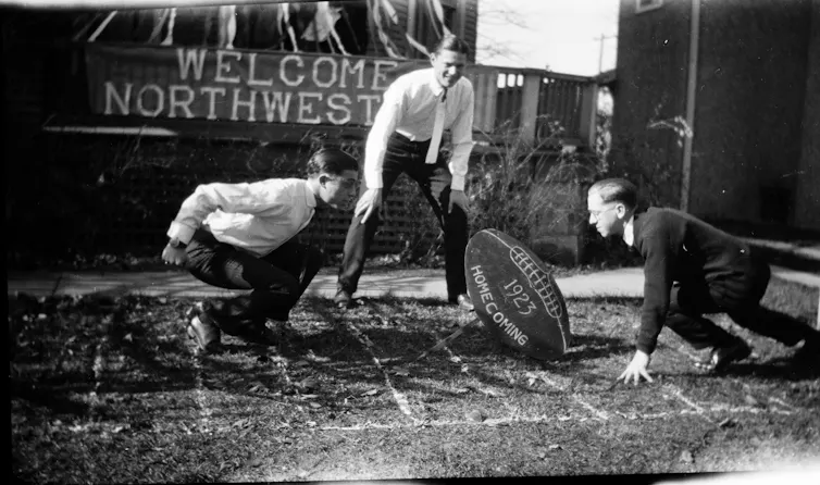 Photo en noir et blanc montrant deux jeunes hommes accroupis de part et d’autre d’un ballon de football surdimensionné, tandis qu’un troisième se tient debout entre eux.